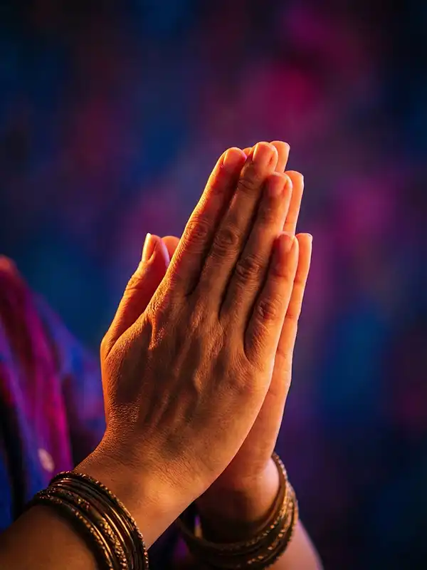 Mãos em Namaskar Mudra com pulseiras, sob iluminação quente de um lado e tons de magenta e azul do outro.
