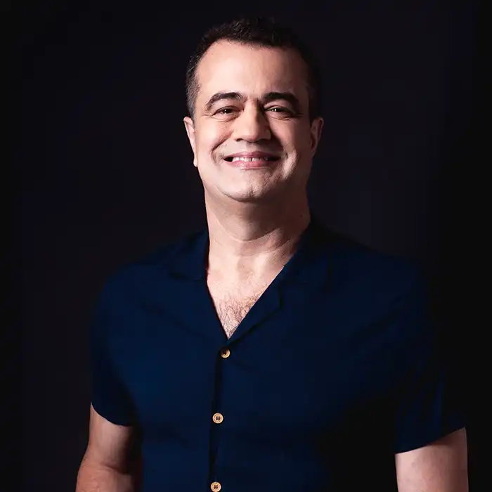 Um retrato profissional de Fábio Nirvano, um homem de pele clara e cabelo curto, sorrindo calorosamente para a câmera. Ele veste uma camisa de manga curta azul marinho com botões de aparência amadeirada, posando contra um fundo preto sólido e escuro.