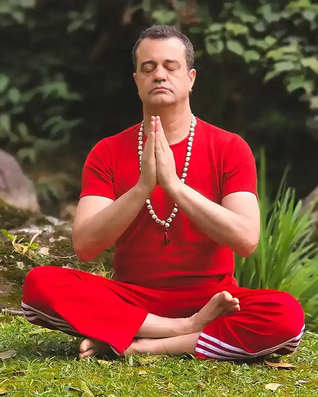 Professor Fábio Nirvano meditando na natureza com as mãos em prece e colar de contas, vestindo vermelho.